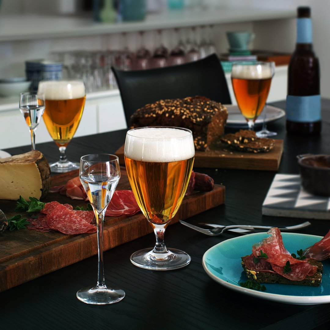 "Elegant dining setting featuring amber beer, clear glassware, vibrant green herbs, and rustic wooden table accents."
