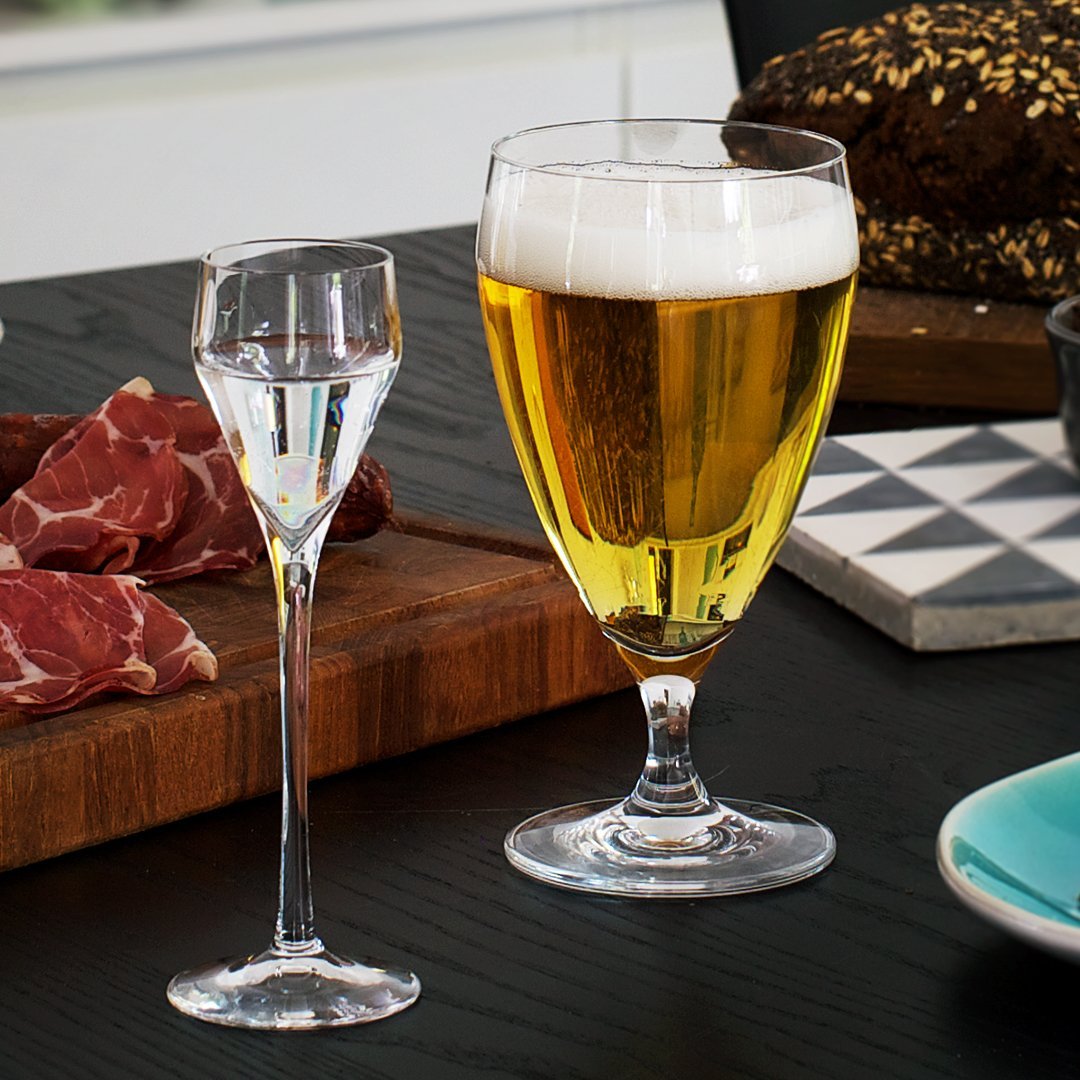Clear glassware displaying golden beer and sparkling liquid, set against a contrasting black table and patterned coaster.