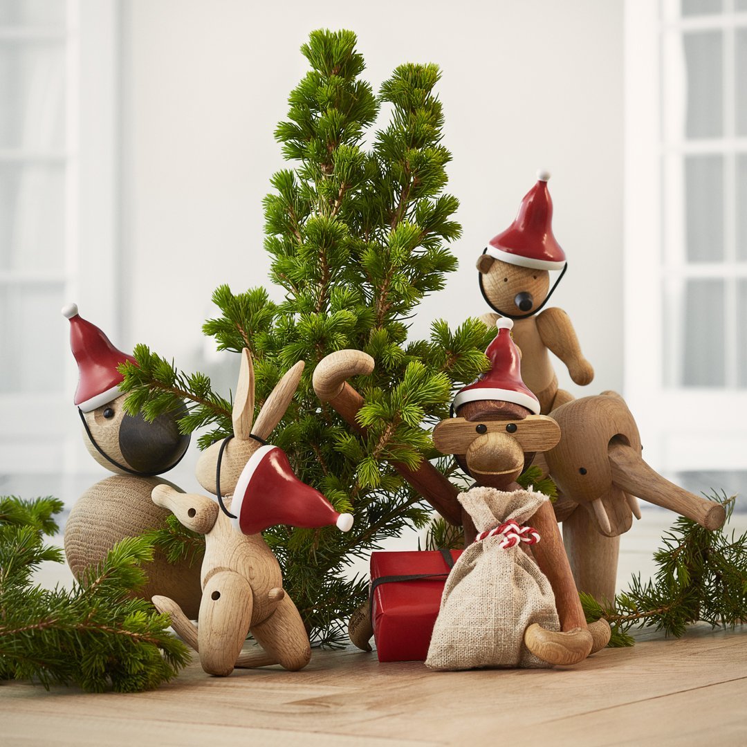 "Wooden animal figurines with red hats and natural tones, surrounded by green foliage and festive decor."