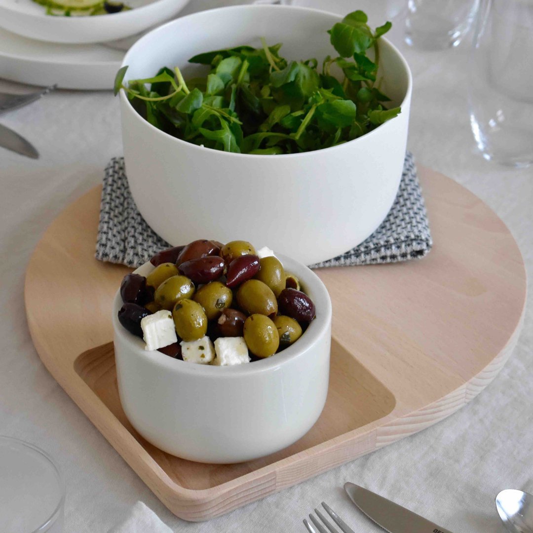 White ceramic bowls on a wooden platter, featuring greens in the larger bowl and olives with feta in the smaller bowl.