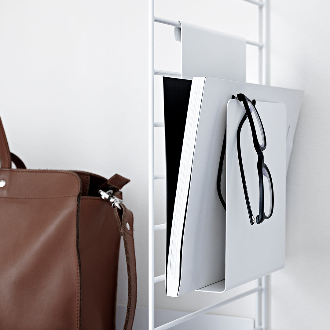 Brown leather bag with black eyeglasses and sleek white notebooks on a white metal shelf, minimalist design.