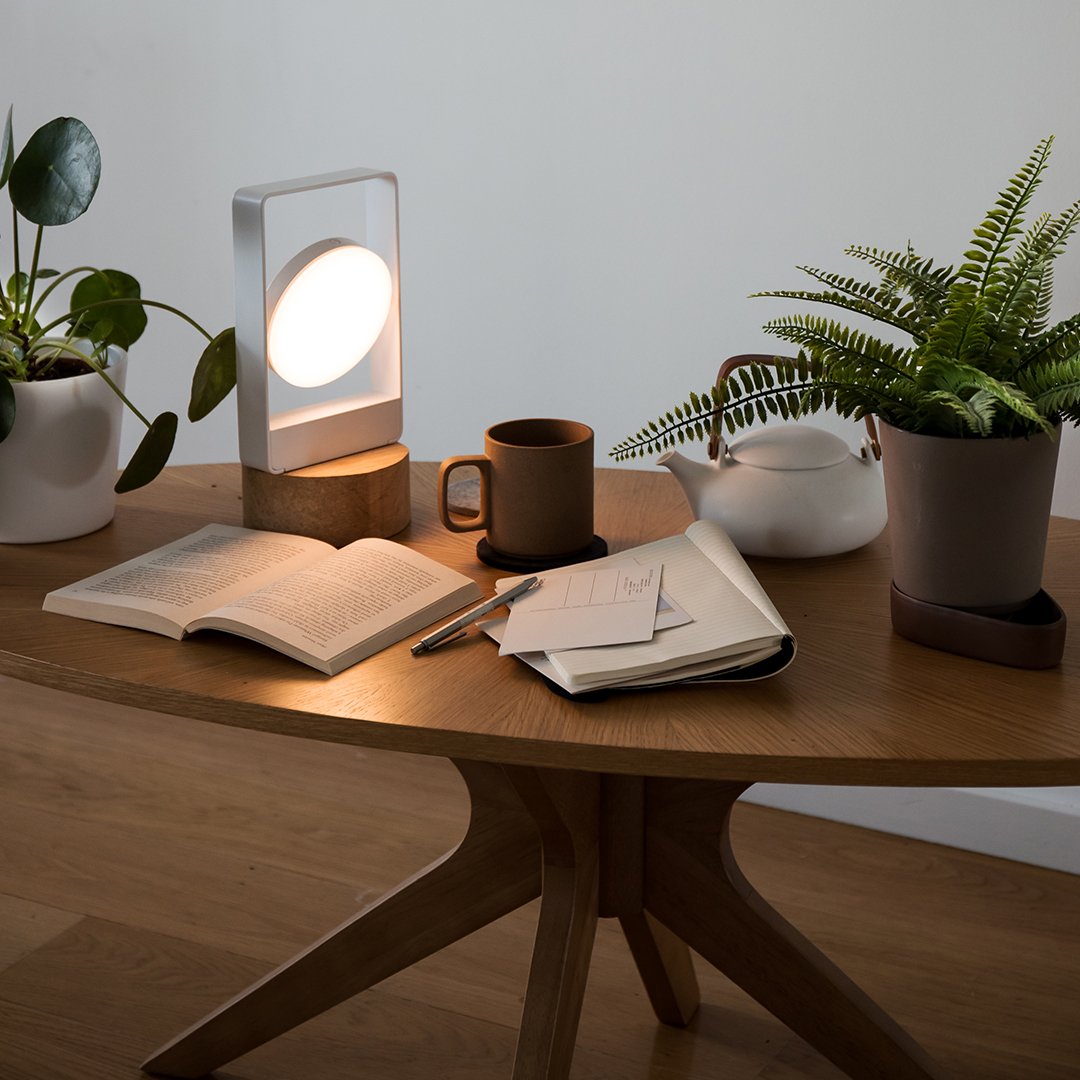 Modern wooden table with white lamp, brown mug, and green plants in neutral pots, featuring soft, warm lighting and natural textures.