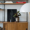Wooden shelf with black frame, featuring white lamp, black artwork, green plant, and accents of red in a modern decor style.