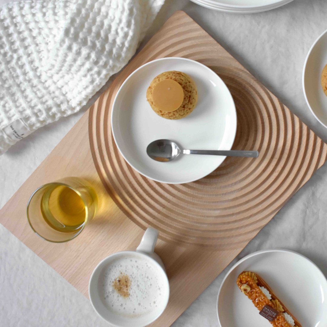 "White plates on a textured wooden tray, featuring a beige cookie, gold drink, and a soft cream-colored towel."