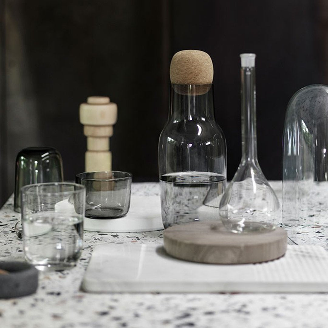 Clear glassware with earthy cork and concrete elements, set against a textured speckled surface. Elegant neutral tones.