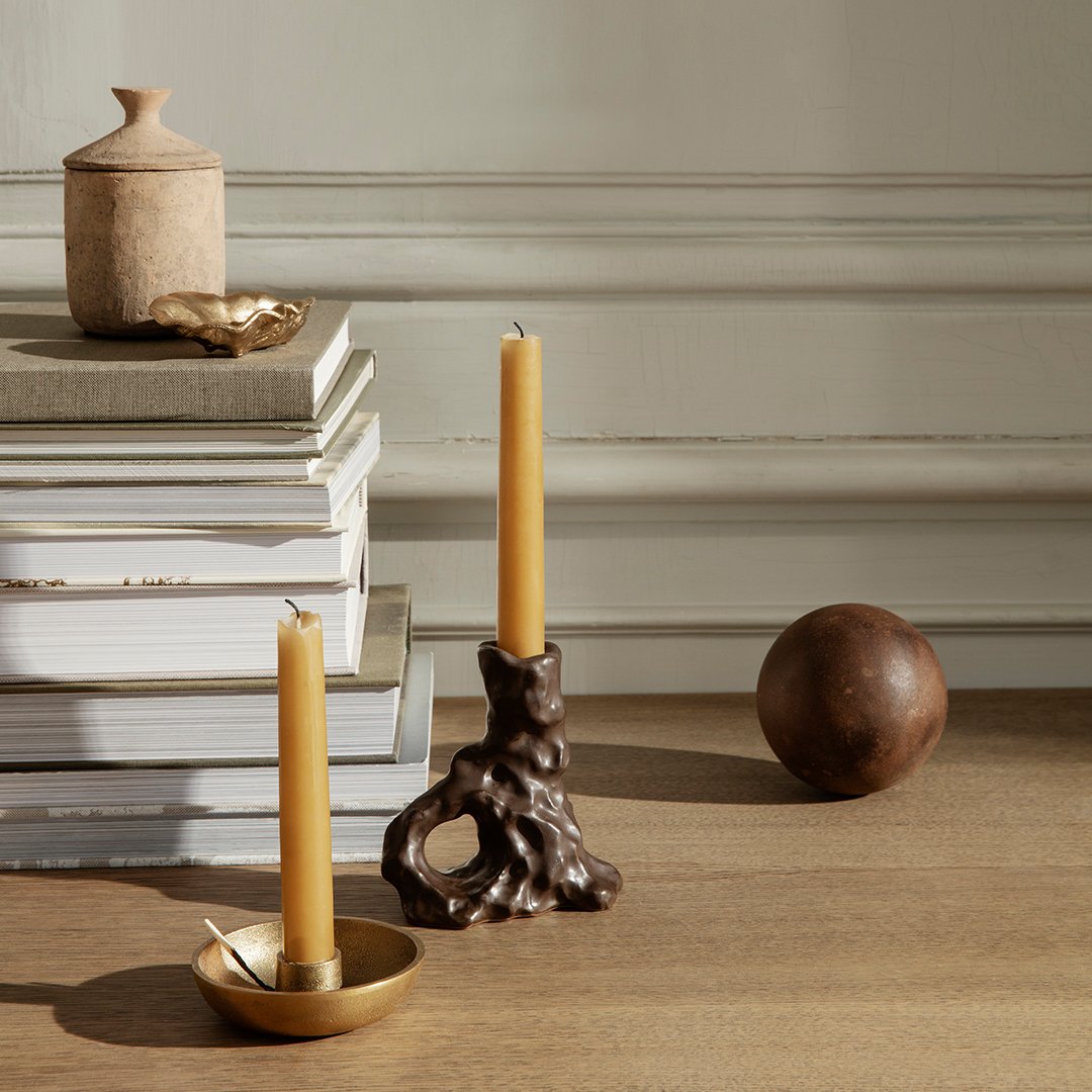 "Rustic brown candles in unique holders, golden accents, and neutral tones against stacked books on a wooden surface."