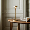 "Modern gold table lamp with a sleek design, set against a neutral-toned wooden table and soft wall backdrop."