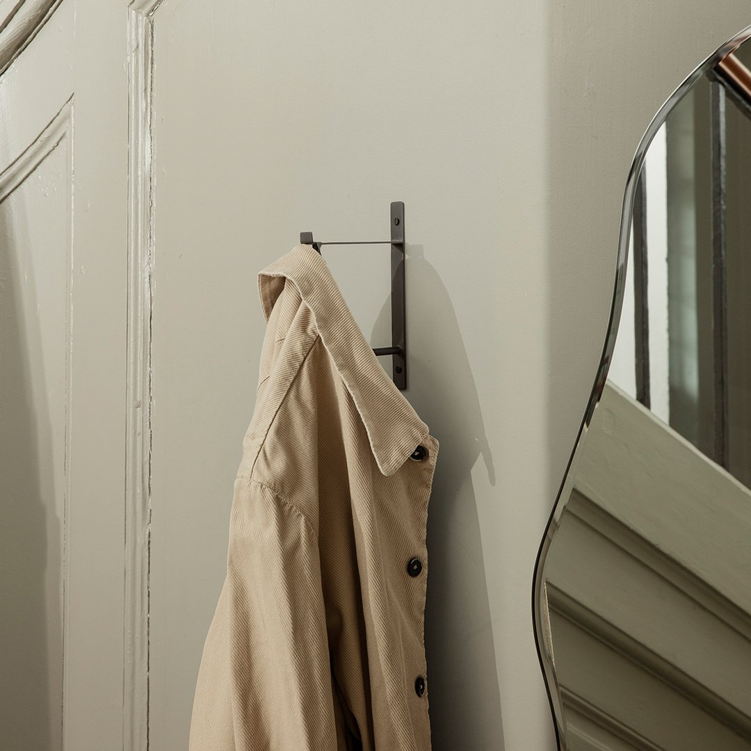 Beige trench coat hanging on a black hook against a neutral wall, with a modern mirror reflecting the space.