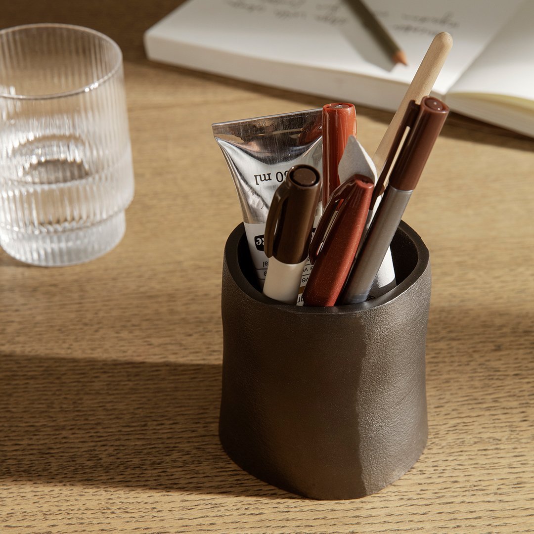 Dark brown ceramic pen holder with glossy finish, featuring a textured design, surrounded by pens and a clear glass.
