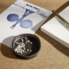 Black round bowl with white pins, set on a wooden surface with soft blue and white artistic prints.