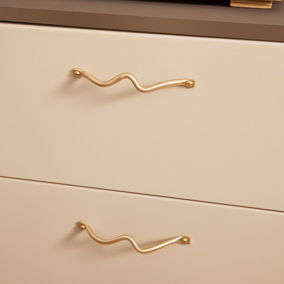 Gold wave-patterned drawer handles on a smooth cream and gray dresser, adding a modern touch to furniture design.