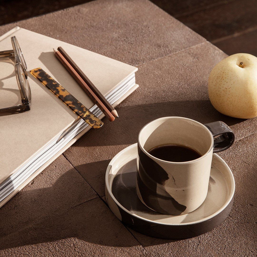 "Beige and black abstract patterned ceramic coffee cup with matching saucer, surrounded by stationery and a pear."