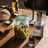 "Clear glass jar with green olives, smoky gray glass bottles, and earthy tones on a wooden table."