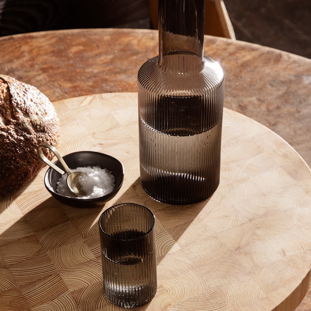 Smoky gray glass carafe and tumbler with vertical ribbed pattern on a wooden table with bread and salt.