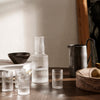 "Clear glass carafe and tumblers on wood table with dark ceramic bowl and pitcher, featuring subtle textured patterns."