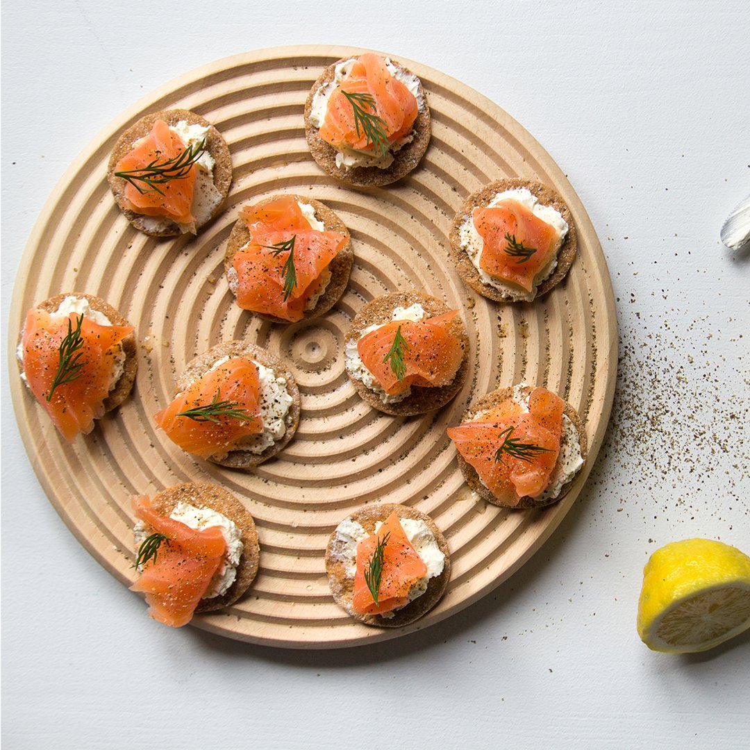 "Round wooden platter with circular grooves showcasing vibrant orange salmon on cream cheese spread, garnished with dill."